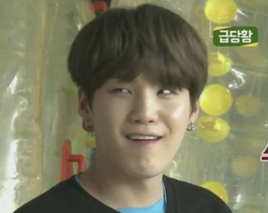 a closeup of bts member suga with a subtle smirk. he has dark hair, light skin, and is wearing a black shirt with a blue collar. the background features a blurred backdrop with yellow orbs and korean text. the shot appears to be taken indoors.