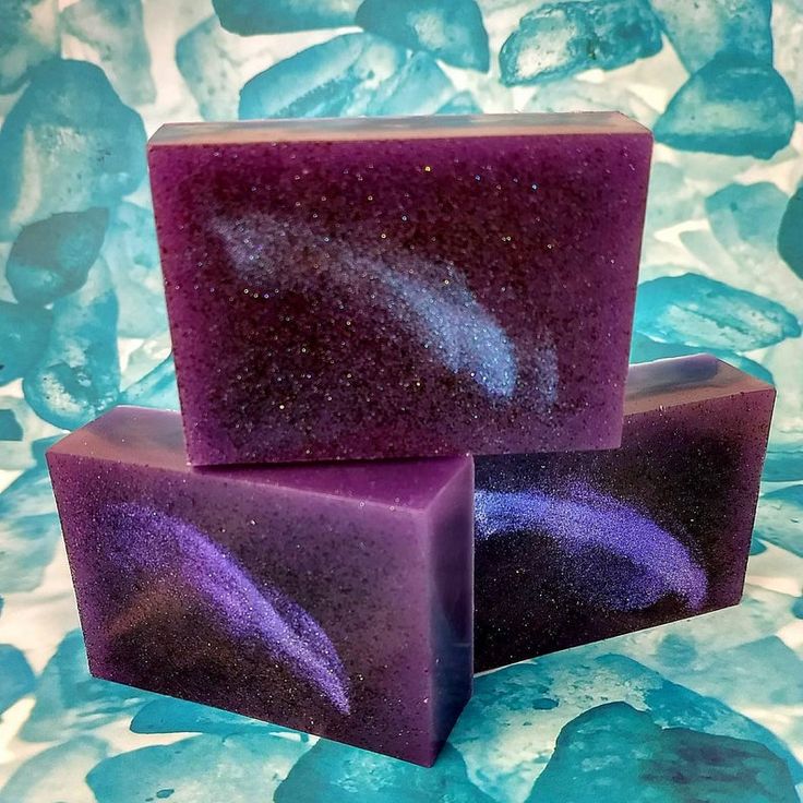 a closeup of three bars of soap, each a deep purple color with a sparkly, galaxylike swirl of blue glitter. the soap is set against a backdrop of a light blue and white textured background. the bars of soap are arranged in a stack with the largest bar on top and the smallest bar on the bottom.