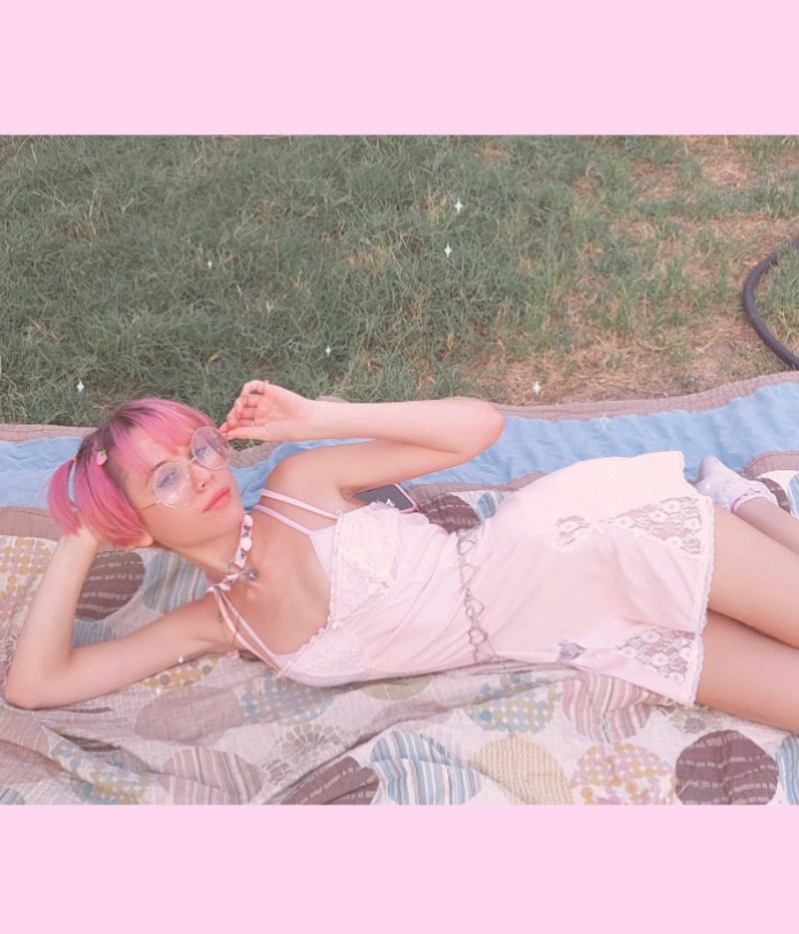 the image shows a young woman with vibrant pink hair lying on a pastelcolored picnic blanket outdoors. she's wearing a delicate, pale pink slip dress with lace detailing, clear round eyeglasses, and a silver body chain.  the overall aesthetic is soft, pastel, and reminiscent of vintage or retro styles with a dreamy, kawaii feel. the scene is sunny and suggests a relaxed, carefree atmosphere.