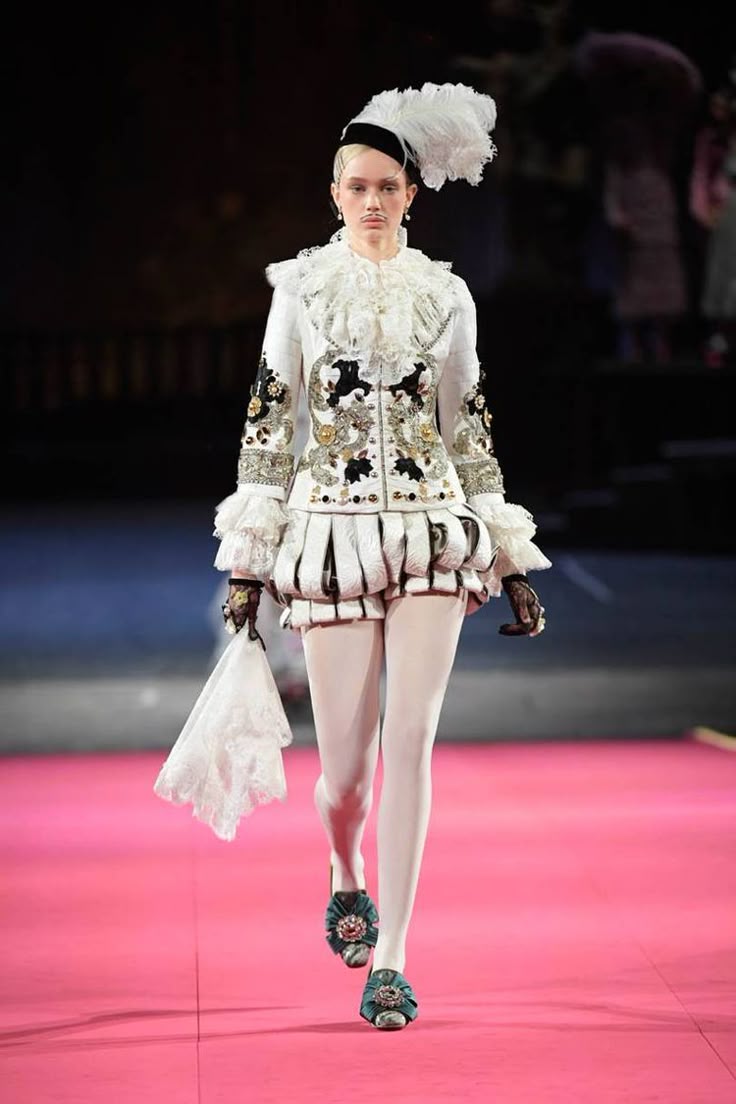 a model walks down a bright pink runway in a white tunic and shorts outfit, white tights, and decorative slipon shoes. the tunic is intricately decorated with black and gold designs, and it has a large, ruffled white collar. the model wears a small black hat with a large white feather. she carries a small, lacy white fan. the background is blurred, with other people visible in the distance.