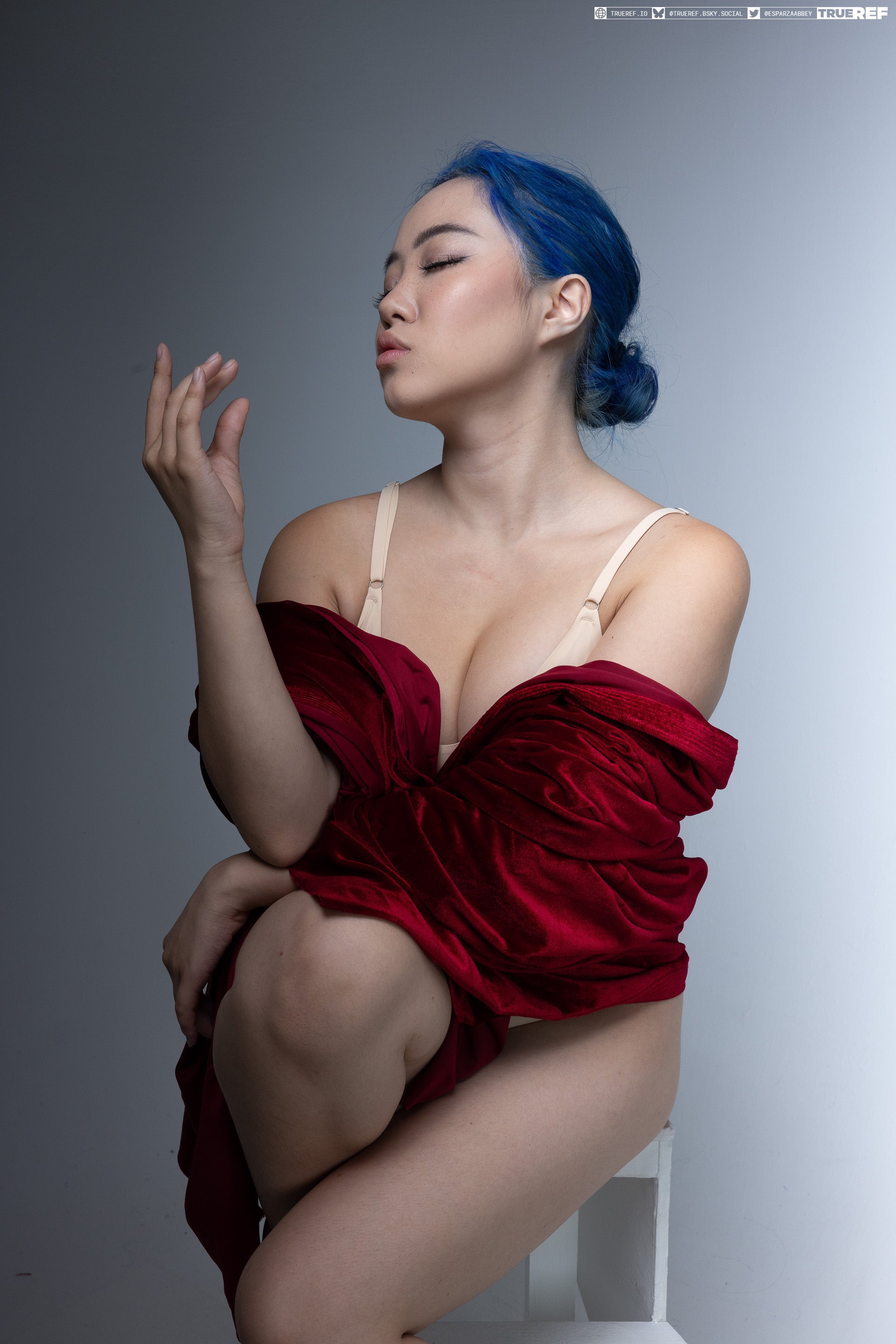 a studio shot of an asian woman with blue hair, seated on a white chair and draped in a red fabric. she is posing suggestively with her eyes closed and a hand raised.