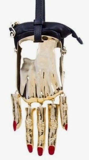 a gold purse shaped like a hand with red nail polish is hanging from a black leather strap. the purse is made of shiny gold material and the fingers are dangling from the bottom. the top of the purse is black leather with a zipper.