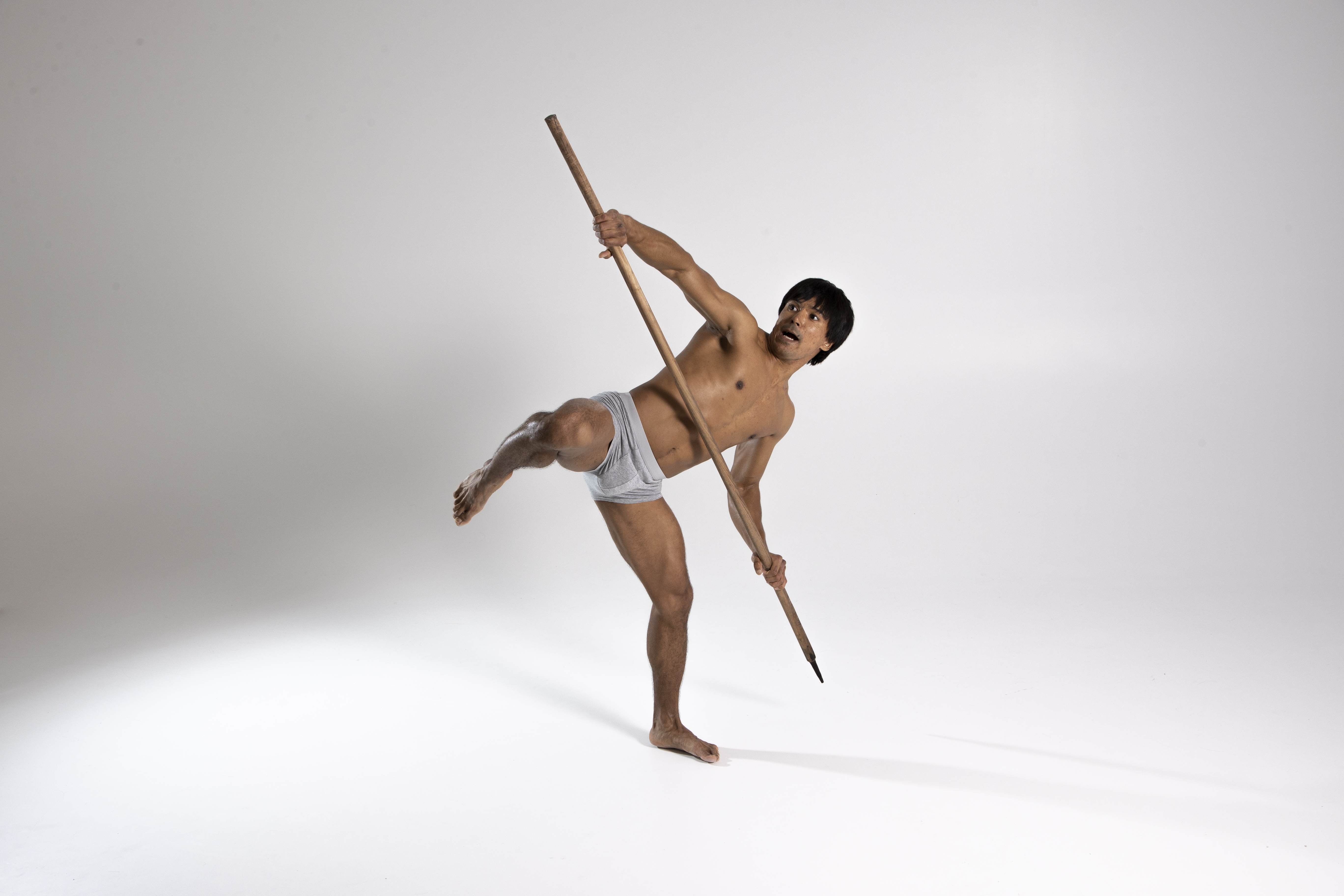 a barefoot man wearing a loincloth is balancing on one leg in a studio shot, holding a spear and looking up.