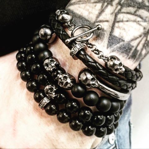 a man's arm is adorned with multiple bracelets. there are two leather bracelets with silver skull accents and chains, along with a stack of beaded bracelets featuring black beads and silver skull accents. the bracelets are worn on a bare arm, with tattoos visible on the man's skin. the lighting is dim, casting shadows on the arm and bracelets.