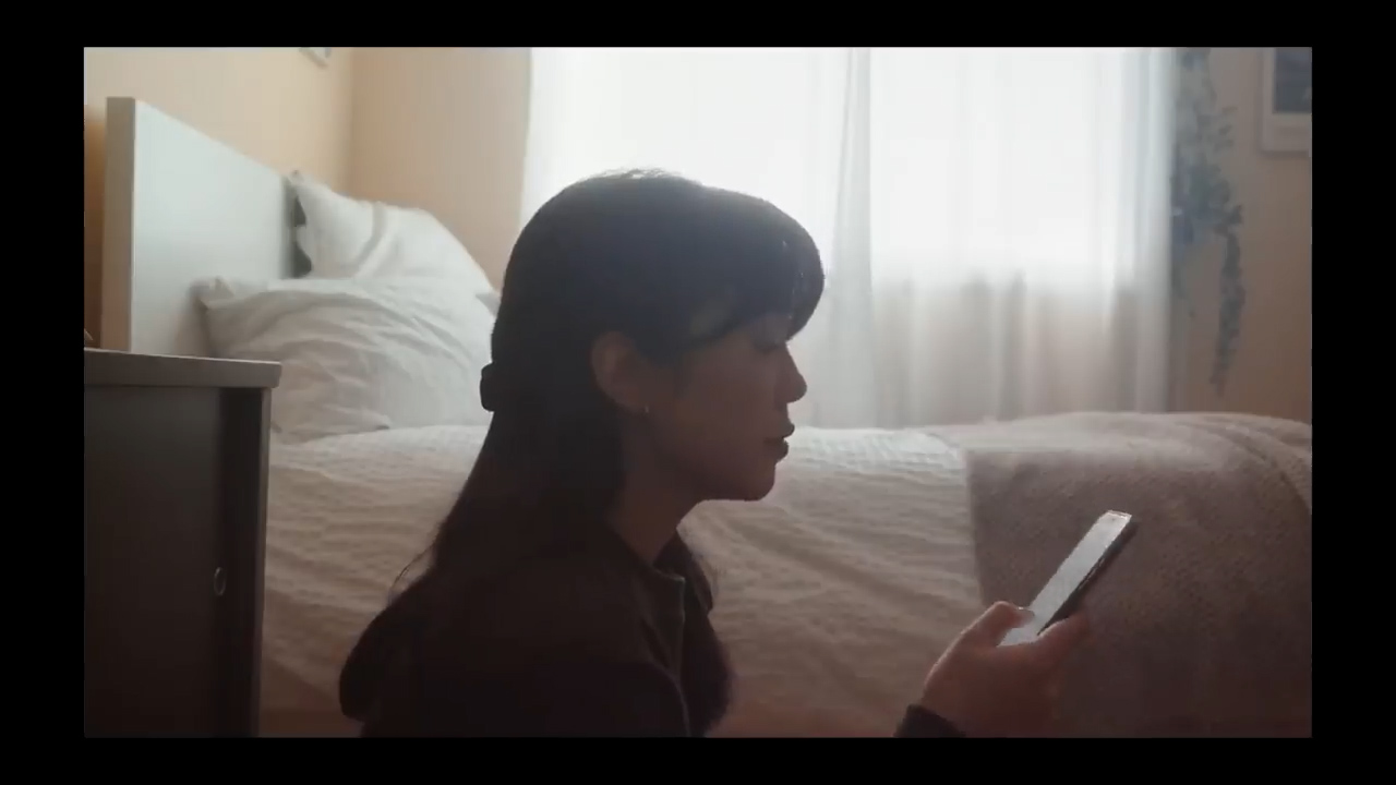 a young woman with dark hair is sitting on the floor of a bedroom, her profile illuminated by soft, natural light streaming in from a window. she's engrossed in her phone, the device held in her hand. the room is simply decorated with a white bed and a dark wooden nightstand in the background.