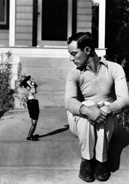 a man is sitting on steps with his legs crossed. he is wearing a collared shirt and pants, and he has a serious expression on his face. the photo is in black and white, and the man is wearing a casual outfit.  a figurine is standing in the background, wearing a hat and suit.  the photo looks like it was taken outdoors.