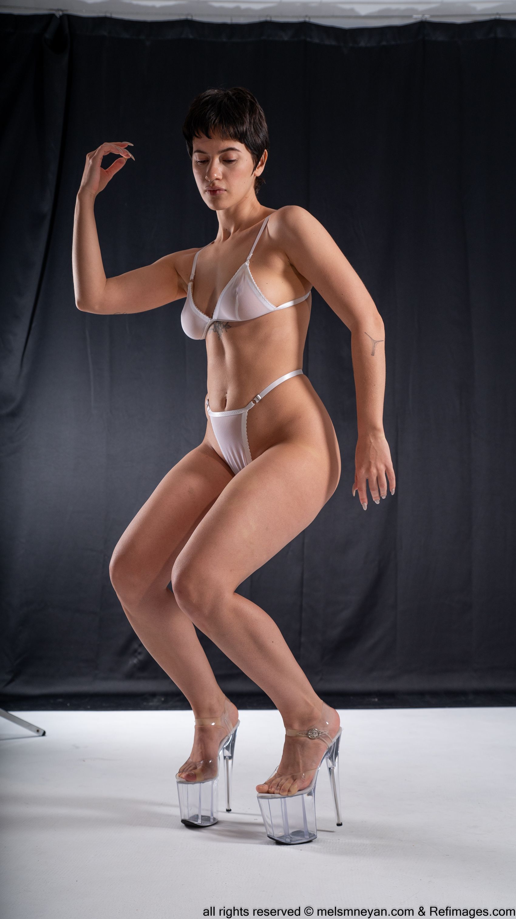 a woman in white lingerie and clear platform heels posing in a studio with a black backdrop.