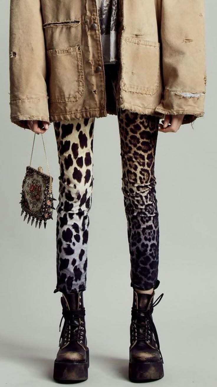 a person stands in a studio with a neutral backdrop, wearing a beige, wornlooking jacket and leopard print jeans that transition from light to dark. their feet are clad in dark brown or black platform boots. in their left hand, they hold a small, ornate purse with spikes. the overall style leans towards grunge or alternative fashion.