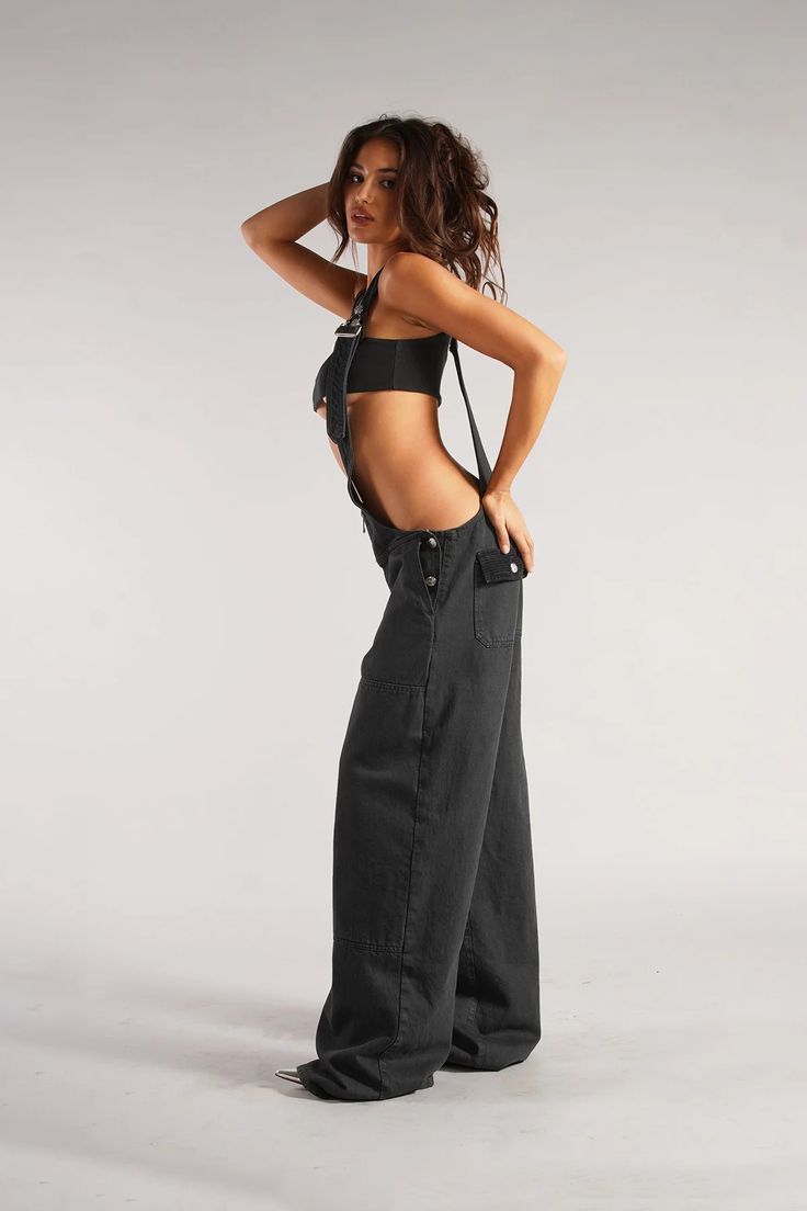 a young woman with long brown hair is posing in a studio setting. she is wearing a pair of black denim overalls with a single strap undone, paired with a black crop top. the image highlights the clothing and the model's physique. the lighting is soft and diffused, creating a gentle contrast and emphasizing the details of the outfit.