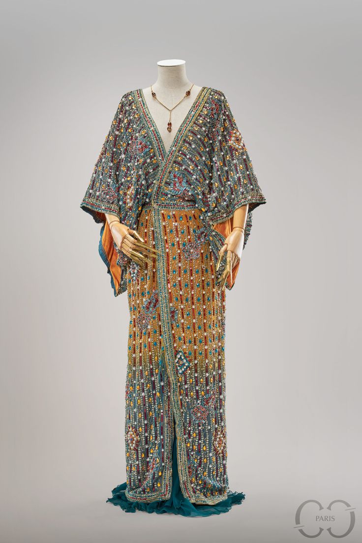 the image features a beaded gown displayed on a mannequin. the gown is elaborately decorated with various beads and embellishments in a range of colors including gold, blue, and red. it has a vneckline and kimonostyle sleeves. a beaded necklace is also visible on the mannequin. the gown appears to be a piece of couture or vintage clothing, possibly from the 1920s based on its style. the fabric is draped and textured with the beadwork. a logo is visible in the bottom right corner.