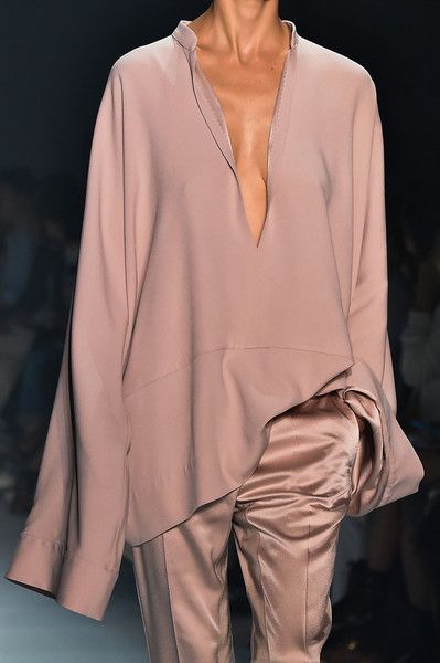 loseup view of a fashion model showcasing an oversized, blushpink blouse with long sleeves and a deep vneck, paired with matching satin trousers. the model is posing on a runway or during a fashion shoot. the overall aesthetic is minimalist and chic, with a focus on the drape and flow of the clothing. the lighting and background are muted, drawing attention to the outfit’s color and texture.