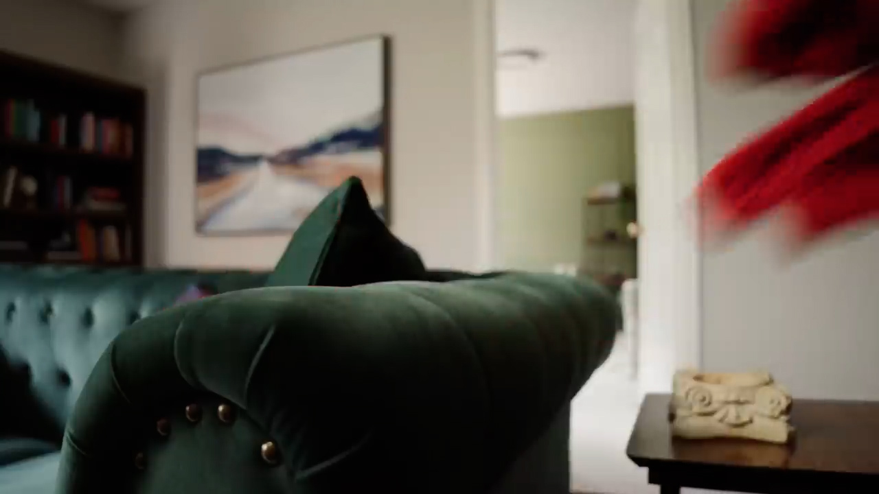 this image shows a stylish living room with a focus on a green velvet sofa. the sofa has tufted detailing and is adorned with a matching pillow. to the left, a bookcase filled with books adds a touch of sophistication. on the wall behind the sofa, a large abstract painting featuring a road leading to mountains adds a touch of artistic flair. the room is welllit with natural light, creating a welcoming and inviting ambiance.