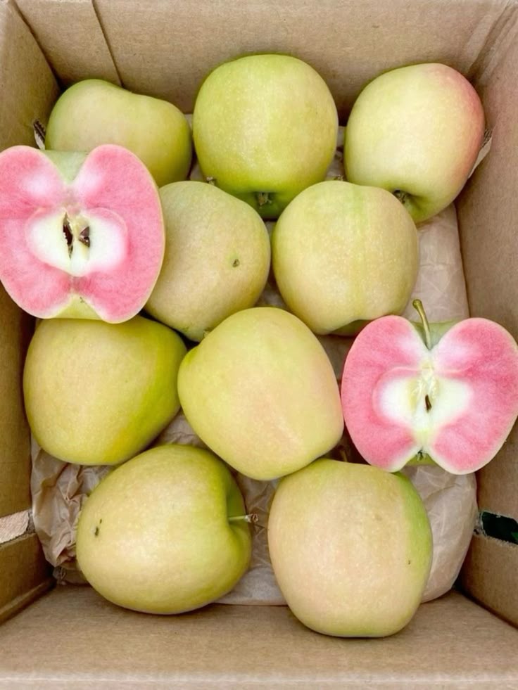 a box of apples, with several whole green apples and two cut apples that reveal pink flesh inside.