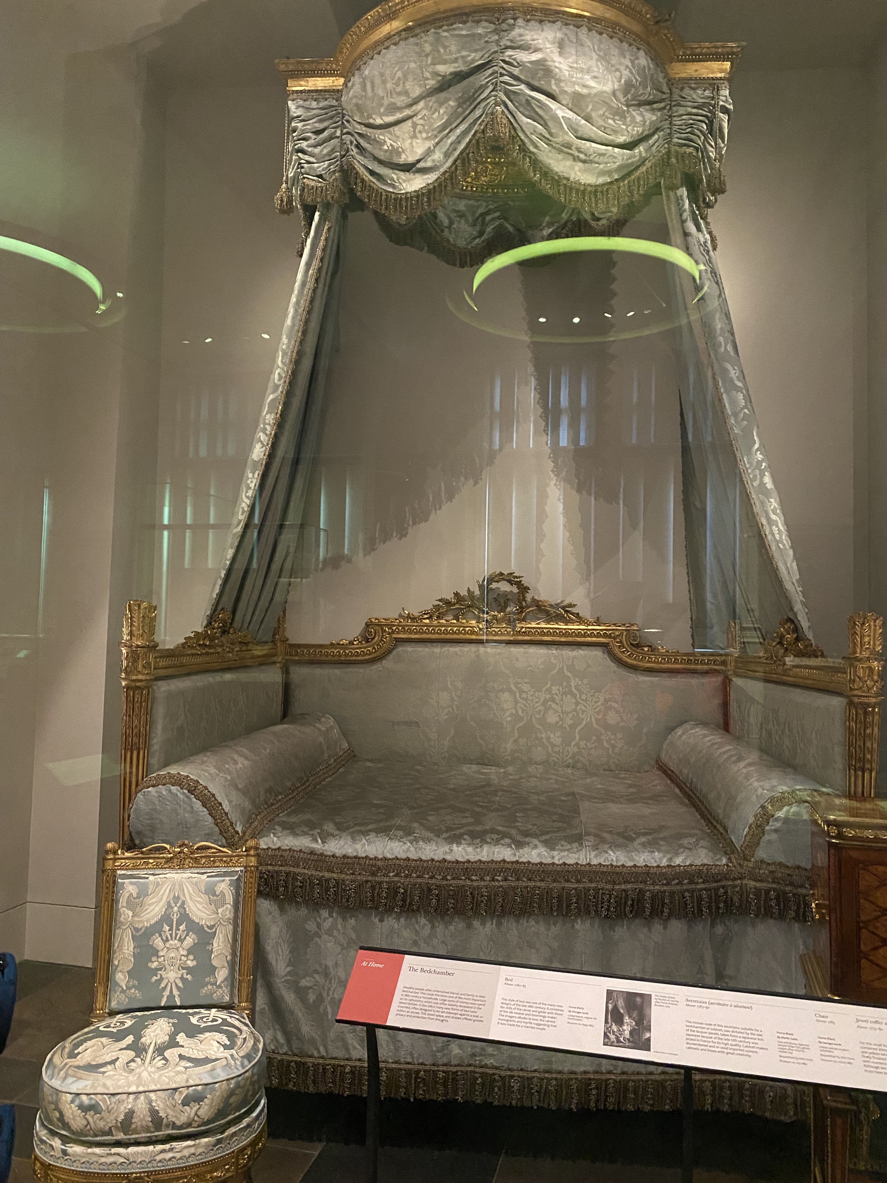 a bed inside a display case at a museum. the bed has a canopy and gold trim. there is also a chair next to the bed. a description card is in front of the furniture.