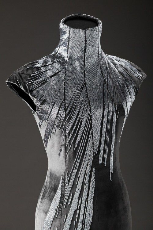 a closeup of a black, highnecked dress adorned with intricate silver beading, creating a stunning and sophisticated design. the beading is arranged in vertical lines that radiate from the center of the neckline, adding a sense of movement and dynamism to the piece. the dress embodies elegance and luxury, showcasing exceptional craftsmanship and artistry.