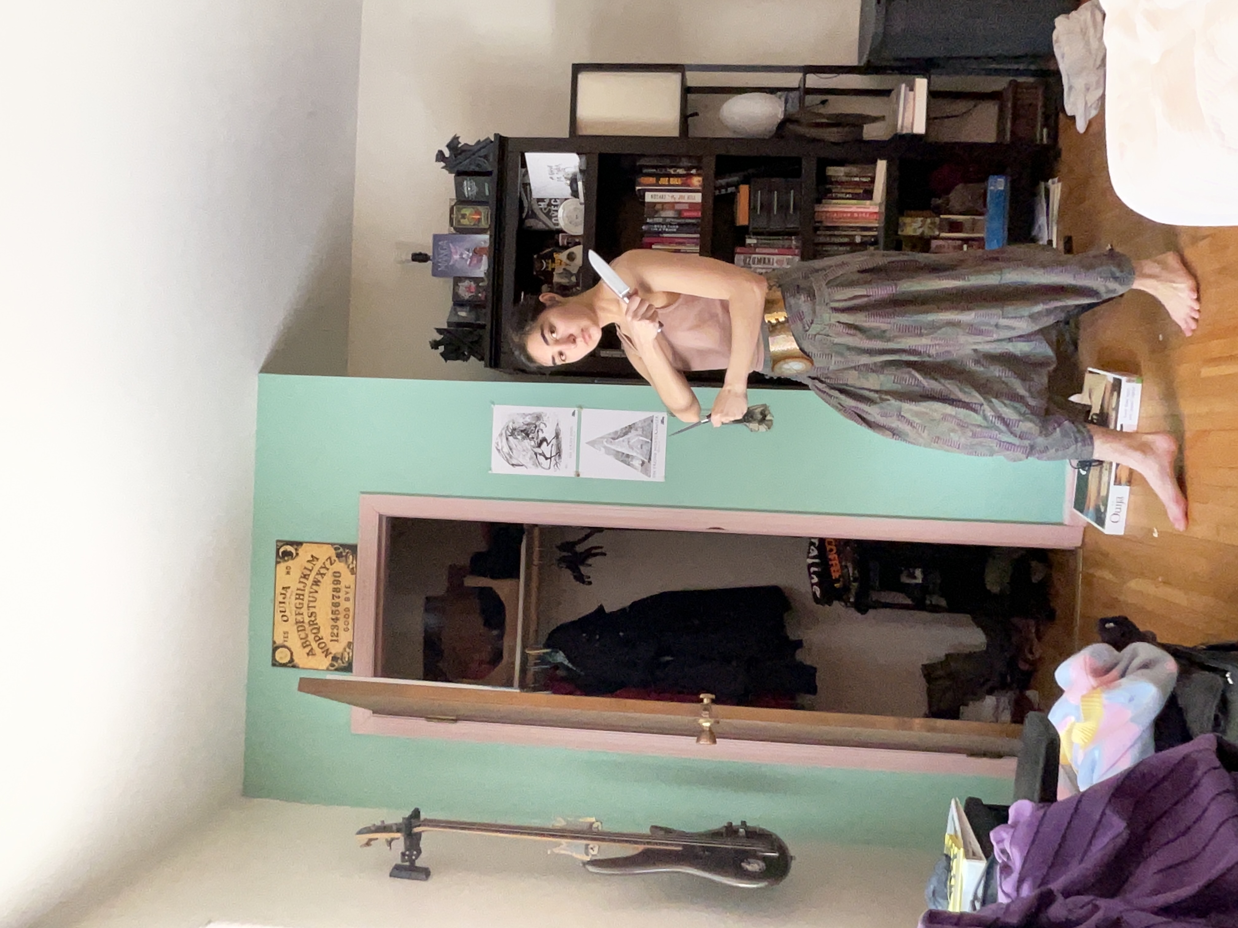 a young woman with long brown hair is standing in her bedroom. she is wearing a light pink tank top and loosefitting, patterned harem pants. she is holding a knife and scissors. the bedroom has pastel green walls, an open closet with clothes hanging inside, and bookshelves filled with books. a ouija board is visible on the wall.  the overall style is eclectic and bohemian.