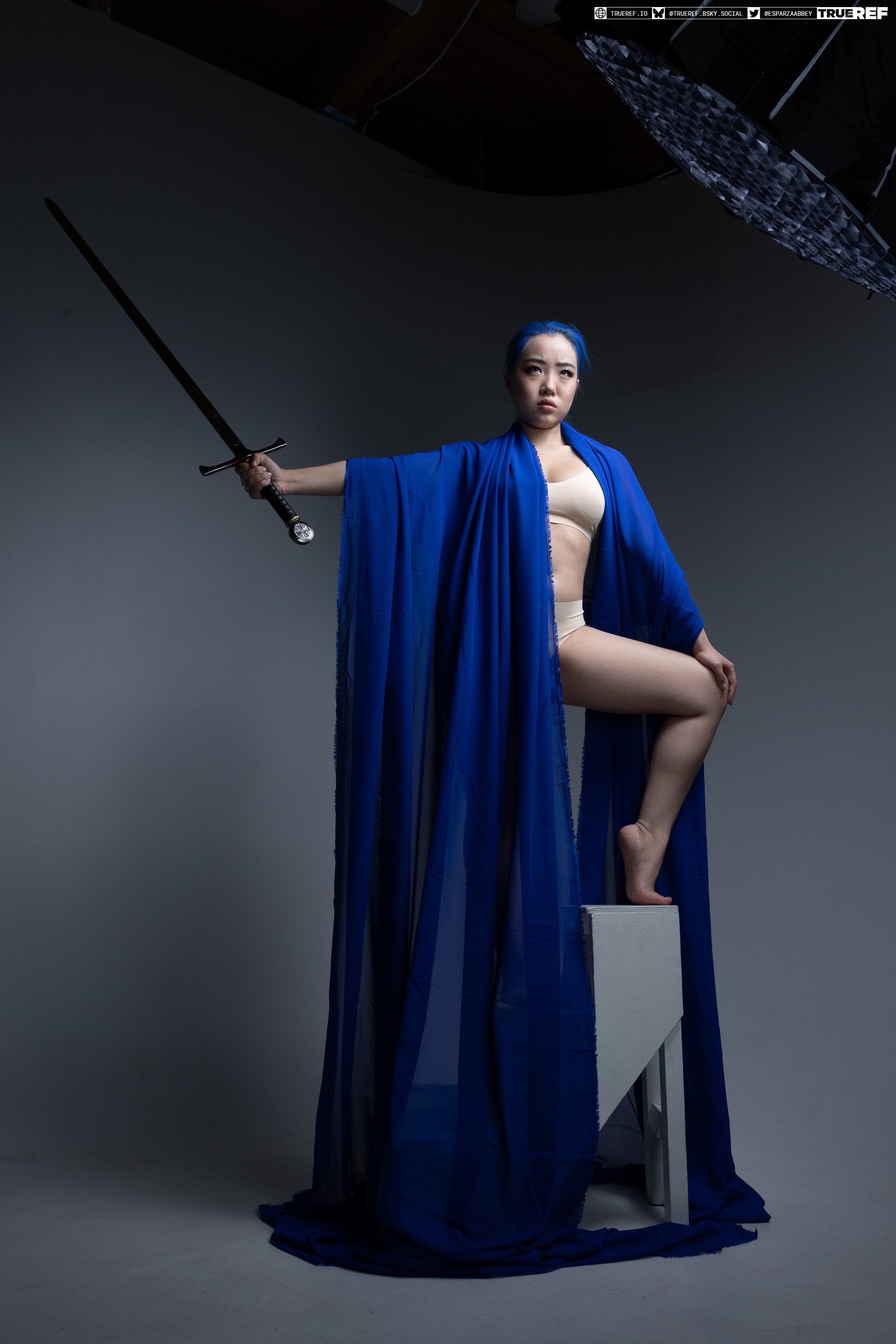 a woman with blue hair is standing on a box in a studio setting. she is wearing a beige bra and underwear, and draped in a long, flowing blue fabric. she holds a sword in her right hand.