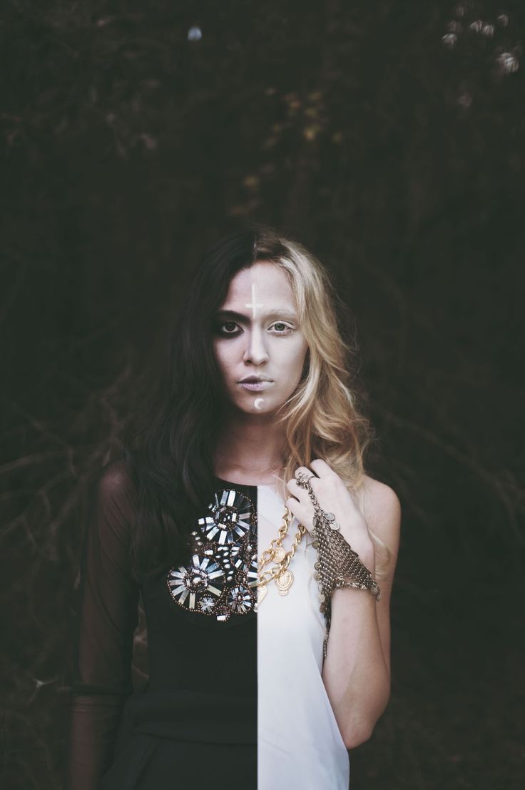 a young woman with dark hair and light skin, adorned with makeup, stands in a dimly lit forest. her hair is styled in a long, flowing manner and parted in the middle, revealing a stark contrast between the dark side and the light side of her face. she wears a black and white dress, accessorized with ornate gold jewelry and a mesh glove on one hand. the background consists of blurred trees and branches, creating a sense of depth and mystery.
