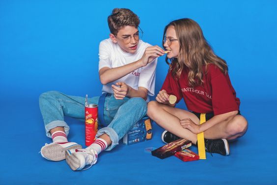 two young adults are sitting on the floor, snacking and smiling at each other, against a blue background. the boy is wearing blue jeans, white tshirt, and sneakers. he is holding a can of soda in his hand and looking at the girl. the girl is wearing red tshirt and jeans, and holding a piece of chocolate in her hand.