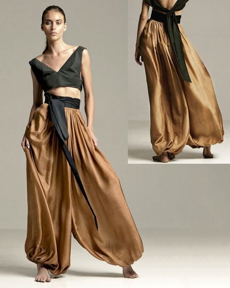 a female model is showcasing a stylish twopiece outfit consisting of a black crop top and voluminous bronze harem pants. the top features a deep vneckline and a wraparound design with a black fabric tie at the waist. the pants are highwaisted, gathered at the ankles, and cinched at the waist with a matching black fabric tie that extends into a long, flowing sash at the back. the model is posing against a neutral background, highlighting the fluidity and elegance of the garment. the studio lighting accentuates the rich texture and drape of the fabric.