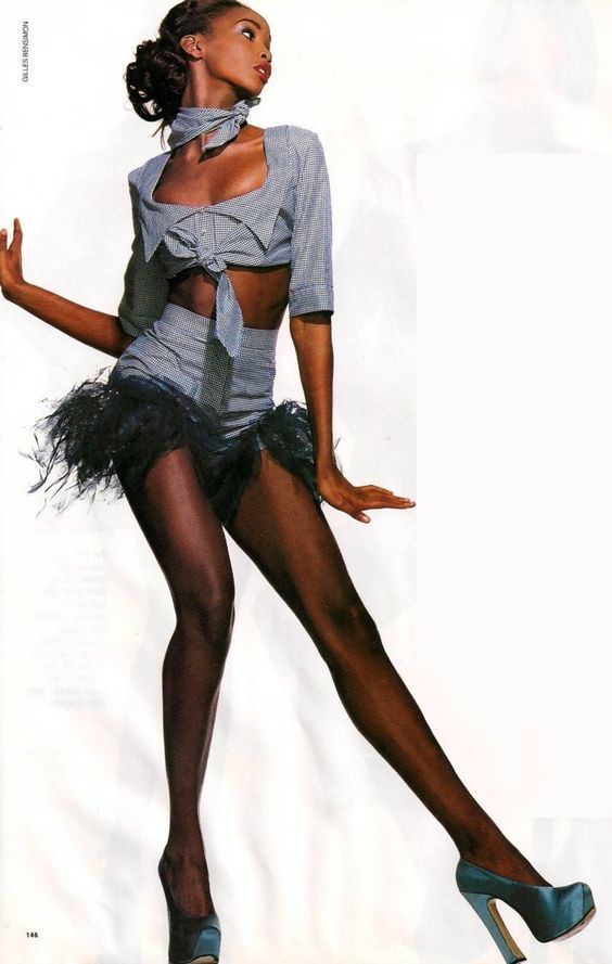 a fashionable woman wearing a blue crop top and skirt paired with high heels and black pantyhose, she is posing in a studio with white background.