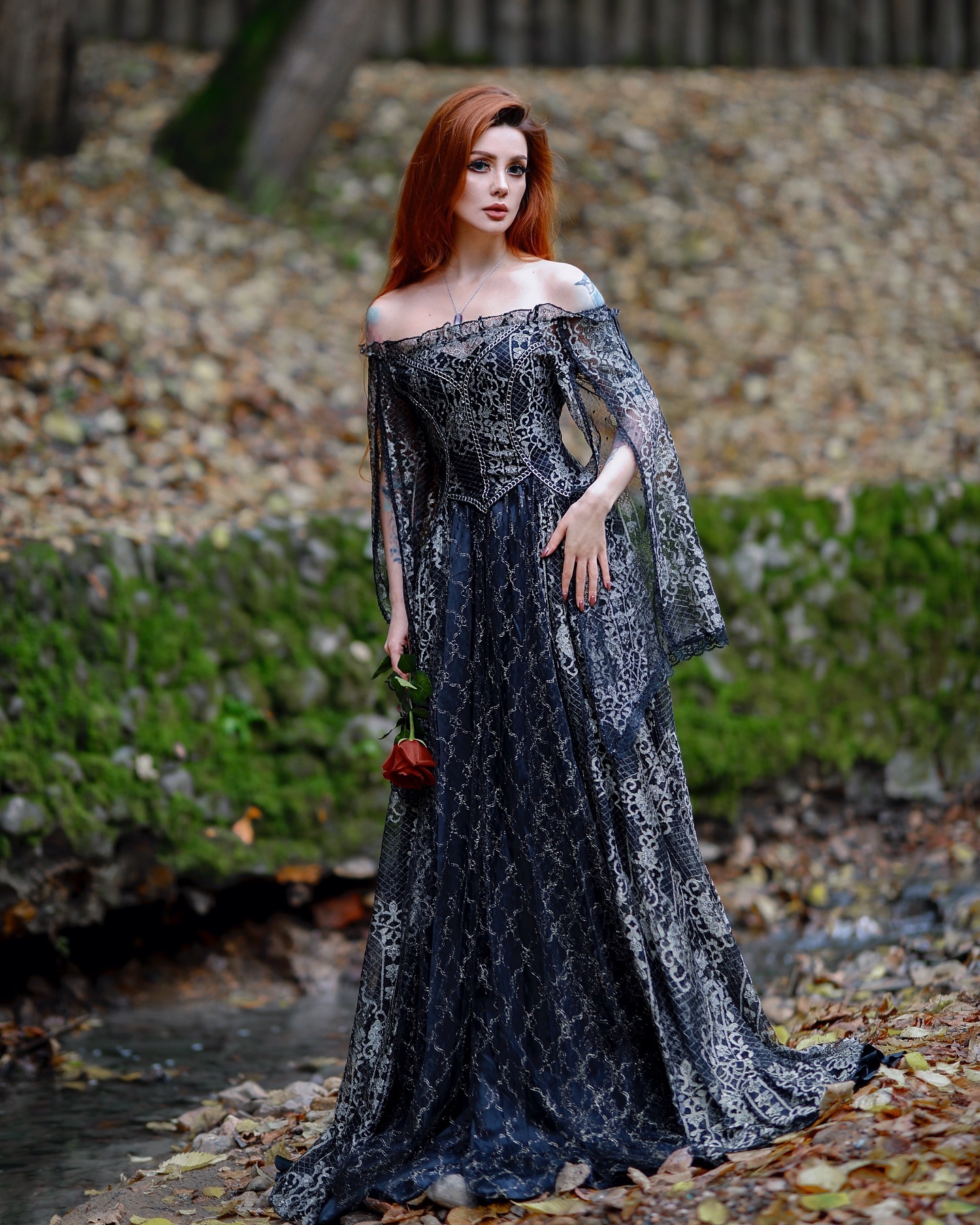a stunning redhead woman, with long flowing hair, stands gracefully by a stream in a forest setting. she wears a long, elegant gothicstyle dress. the dress is predominantly dark navy blue/black with intricate silver lace detailing and offtheshoulder sleeves. the dress has a fitted bodice and a flowing skirt that pools around her ankles.  she holds a single red rose in her hand. the autumnal backdrop of fallen leaves and the natural setting enhances the romantic and slightly mysterious ambiance of the portrait.  the overall mood of the image is dark, romantic, and evocative of a fairytale or fantasy setting.
