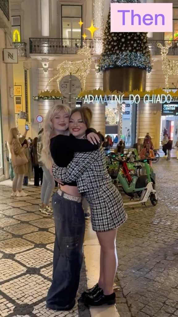 two women embrace each other in front of a building adorned with christmas decorations. one woman is wearing a black crop top and jeans while the other is wearing a black and white patterned coat. a christmas tree and other decorations are visible in the background.