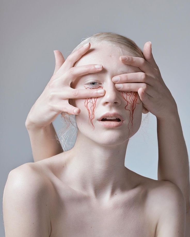 a woman with pale skin, a delicate face, and red tears running down her cheeks, holds her hands over her eyes in a portrait. her expression is one of sadness and pain. the image is stark and dramatic, with a white background that accentuates the woman's features. the red tears stand out against her white skin, creating a striking contrast that draws the viewer's attention. the image is both beautiful and unsettling, leaving the viewer with a sense of unease and wonder.