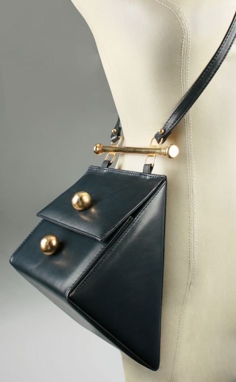 a closeup studio shot presents a structured, triangular leather handbag in a dark color, potentially black or navy, hanging from a mannequin's shoulder. the bag features a unique geometric design with a flap closure, adorned with two prominent goldcolored spherical accents. a matching strap, also made of leather, crosses over the mannequin's shoulder and connects to the bag via a horizontal gold bar with white or lightcolored ends, adding an element of visual interest and structural support. the background is a simple, neutral gray, drawing full attention to the bag and its design. the lighting is wellbalanced, highlighting the texture of the leather and the shine of the gold accents.