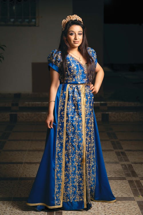 a fulllength portrait of a young woman elegantly dressed in a vibrant blue moroccan caftan (takchita). the caftan features intricate gold embroidery along the bodice and vertical panels, adding a luxurious touch.  she wears a gold headpiece or tiara. the setting appears to be an interior with a tiled floor.  the overall image projects an air of sophistication and showcases the rich details of the traditional garment.