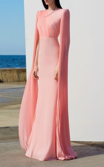 a full shot of a woman standing against a blurredout background of an ocean and a walkway, wearing a long, elegant light pink dress. the dress has pleated cape sleeves and a fitted bodice. the overall style is formal and sophisticated. the model has dark hair and her posture is serene.
