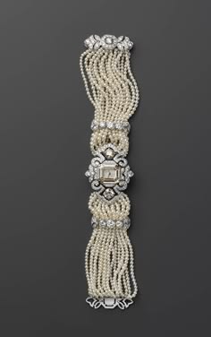 a closeup image of a luxurious pearl and diamond watch bracelet. the watch face is square with a delicate diamond frame, and the bracelet is comprised of multiple strands of pearls and diamonds. the watch is sitting on a grey background.