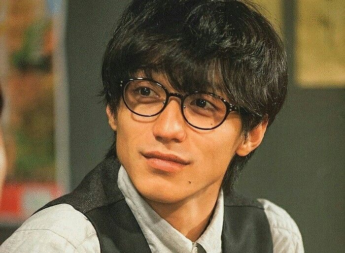 the image is a closeup portrait of kento yamazaki, a wellknown japanese actor. he's wearing roundframed glasses, his dark hair styled with bangs falling across his forehead.  he has a soft, slightly serious yet pleasant expression, almost a subtle smile. he's dressed casually in a white shirt with a darkcolored vest. the lighting is soft and warm, giving the image a cinematic feel, suggesting it might be a still from a film or television drama.  the background is blurred, drawing focus to yamazaki.