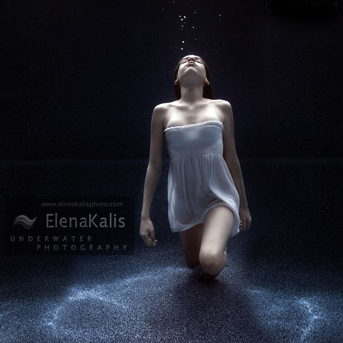 the image showcases a young woman submerged in dark water. she is elegantly dressed in a white, grecianinspired dress. her head is tilted back, her eyes closed, and a stream of bubbles rises from her mouth, creating an ethereal and serene atmosphere. the lighting is lowkey, coming from a source above her, which casts a soft spotlight on her figure and the rising bubbles. the perspective is low, emphasizing her vulnerability and the vastness of the surrounding water.  the overall mood is one of peace, tranquility, and surrender.