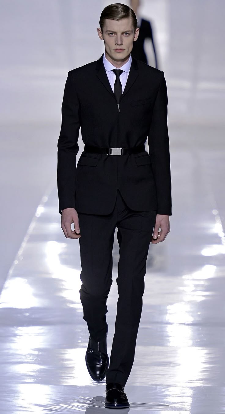 a man is walking down a runway wearing a black suit with a black tie and a black belt. he is also wearing black dress shoes. the background is a white, reflective surface.