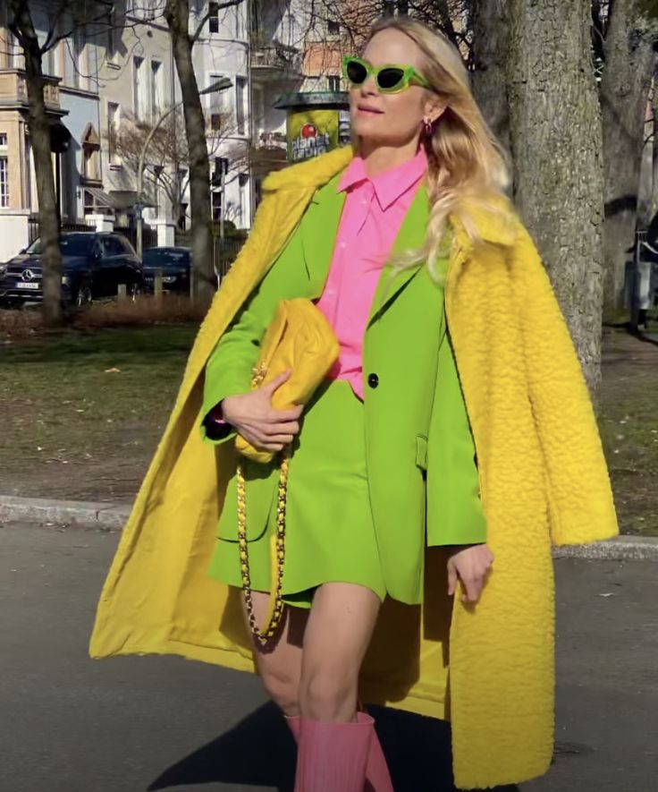 a woman with blonde hair wearing bright colors is standing on a street. she is wearing green sunglasses, a pink shirt, a green blazer and skirt, pink boots, and carrying a yellow handbag. a yellow coat is draped over her shoulders. the background includes trees, buildings, and parked cars.