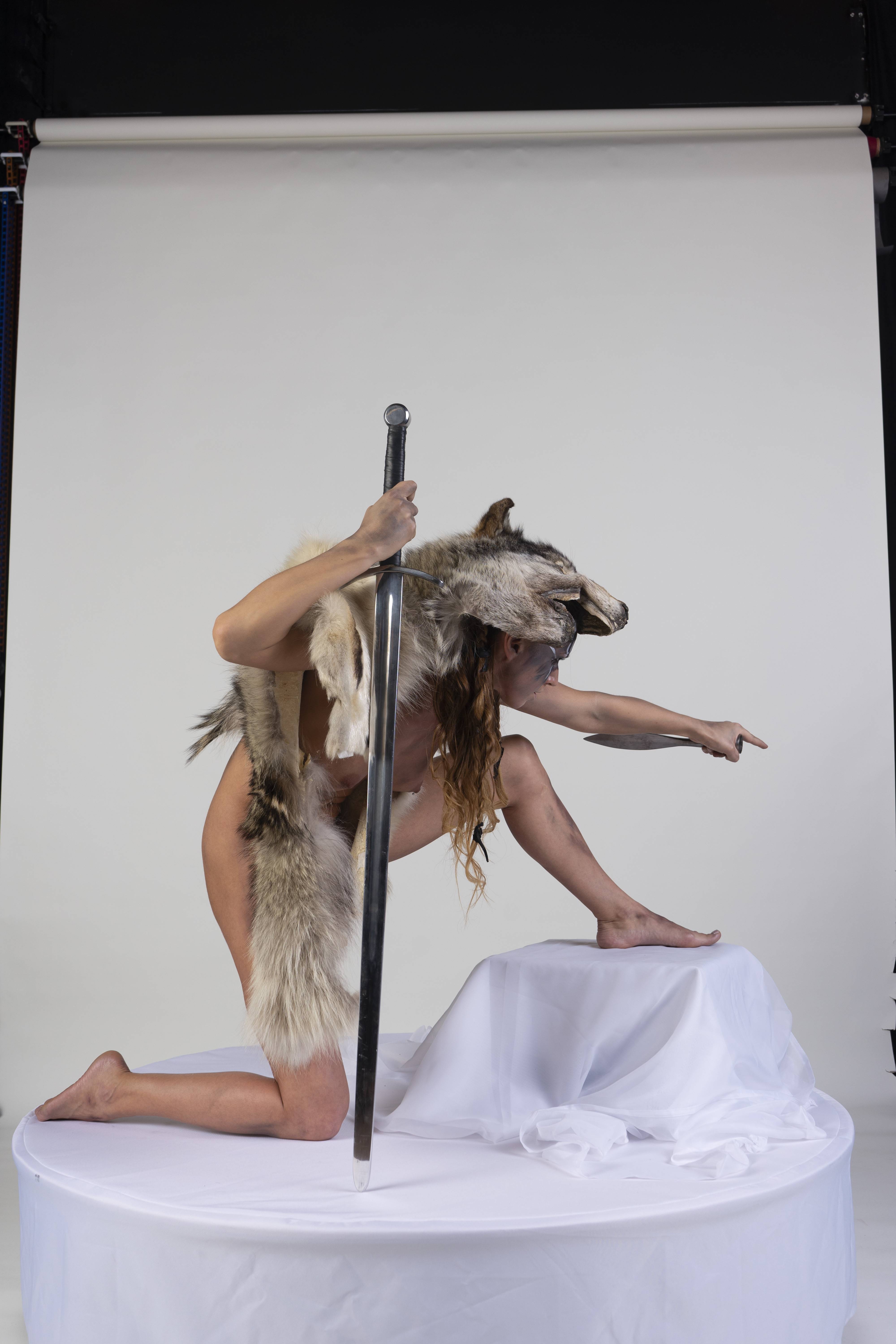 a woman wearing a wolf headpiece and fur shawl is kneeling on a white platform in a studio setting. she is holding a sword and pointing with her other hand. the background is a plain white backdrop.