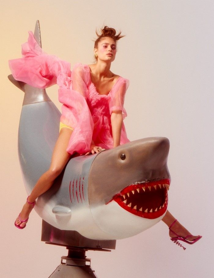 a woman in a pink dress and pink shoes is sitting on a shark's back. she is  posing for the camera in a studio setting. she looks directly at the camera with a serious expression on her face.