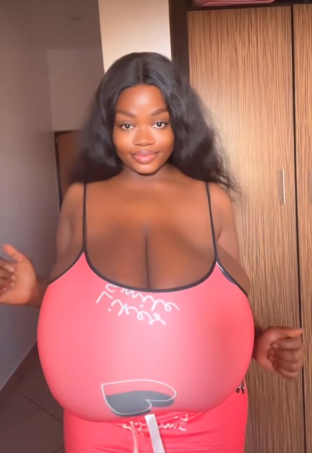 a darkskinned woman with large breasts stands in a bedroom wearing a red tank top and matching pajama bottoms. she is smiling and looking at the camera. the background shows a wooden closet. the image appears to be a selfie or a photo taken by someone close to her. the overall tone suggests body positivity and selfacceptance.