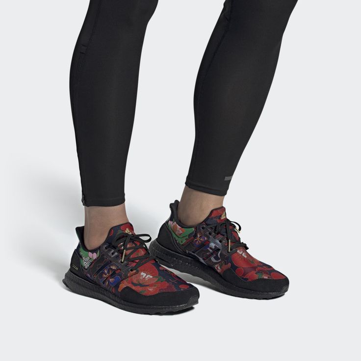a closeup of a person wearing a pair of adidas ultra boost shoes with a floral print. the shoes are black with red, green, and yellow flowers. the person is wearing black leggings. the shoes are on a white background.