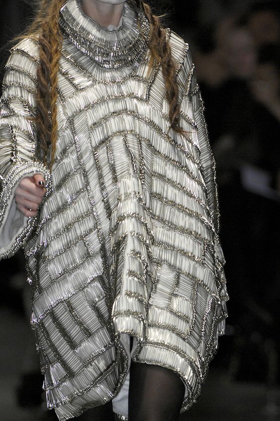 the image shows a model on a fashion runway wearing an elaborate dress constructed entirely from safety pins. the dress has a geometric pattern and a high, ruffled collar. the model has long braids. the overall effect is visually striking and avantgarde, characteristic of high fashion shows and designers such as alexander mcqueen. the background is dark and blurry, suggesting a fashion show setting.
