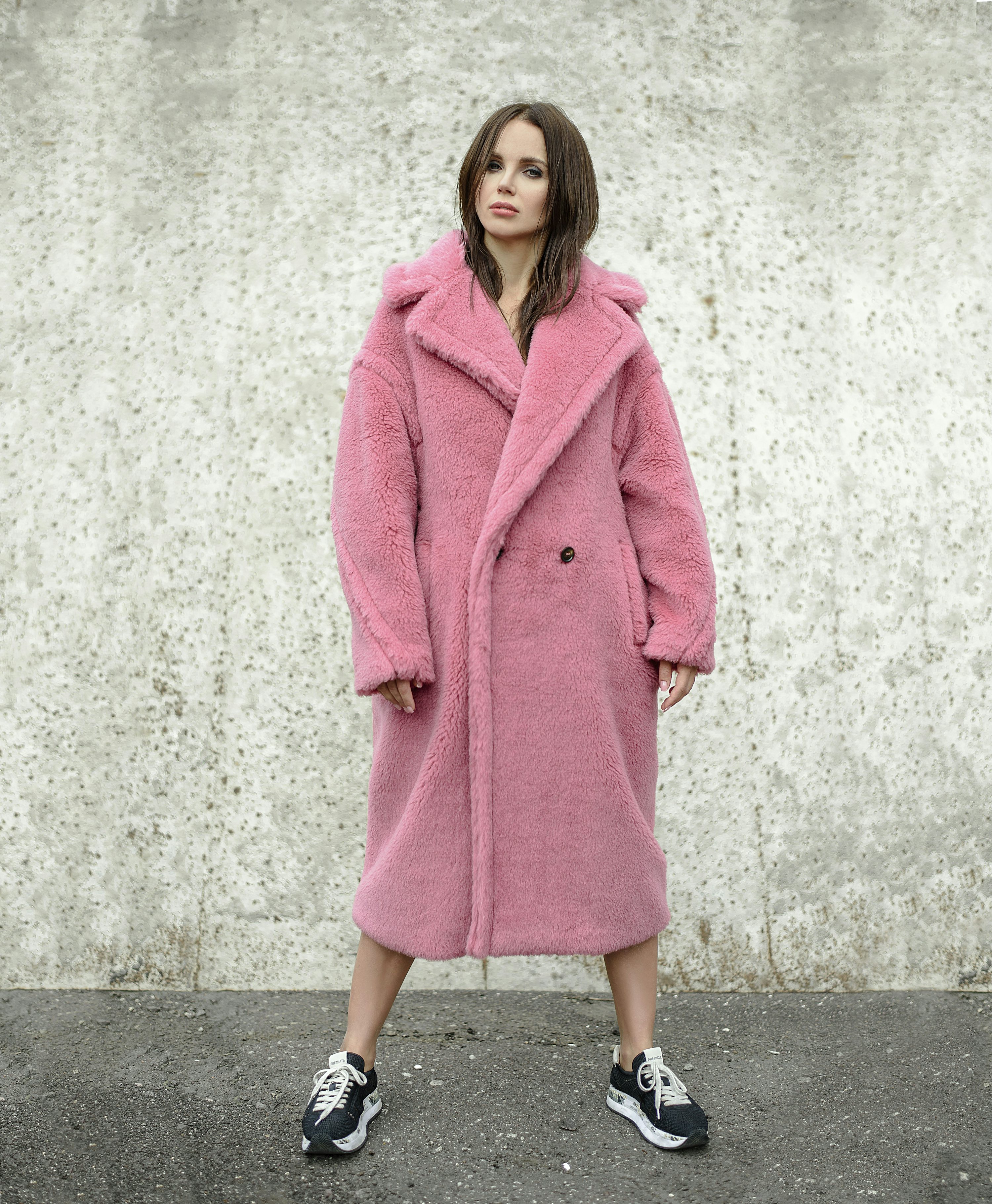 a fulllength shot of a woman standing against a concrete wall. she is wearing a long, oversized, light pink teddy coat, black sneakers, and has long brown hair. the coat is doublebreasted and has a relaxed fit.  the overall style is casual and chic.
