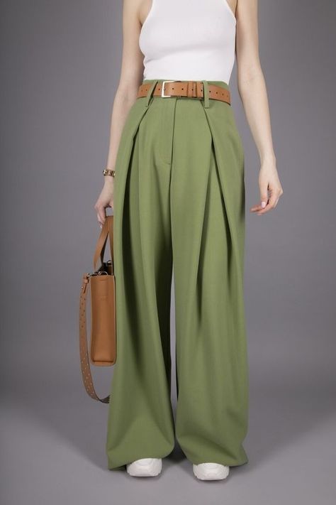 a woman in a white tank top, brown belt, wideleg green pants, and a brown bag stands against a gray background.
