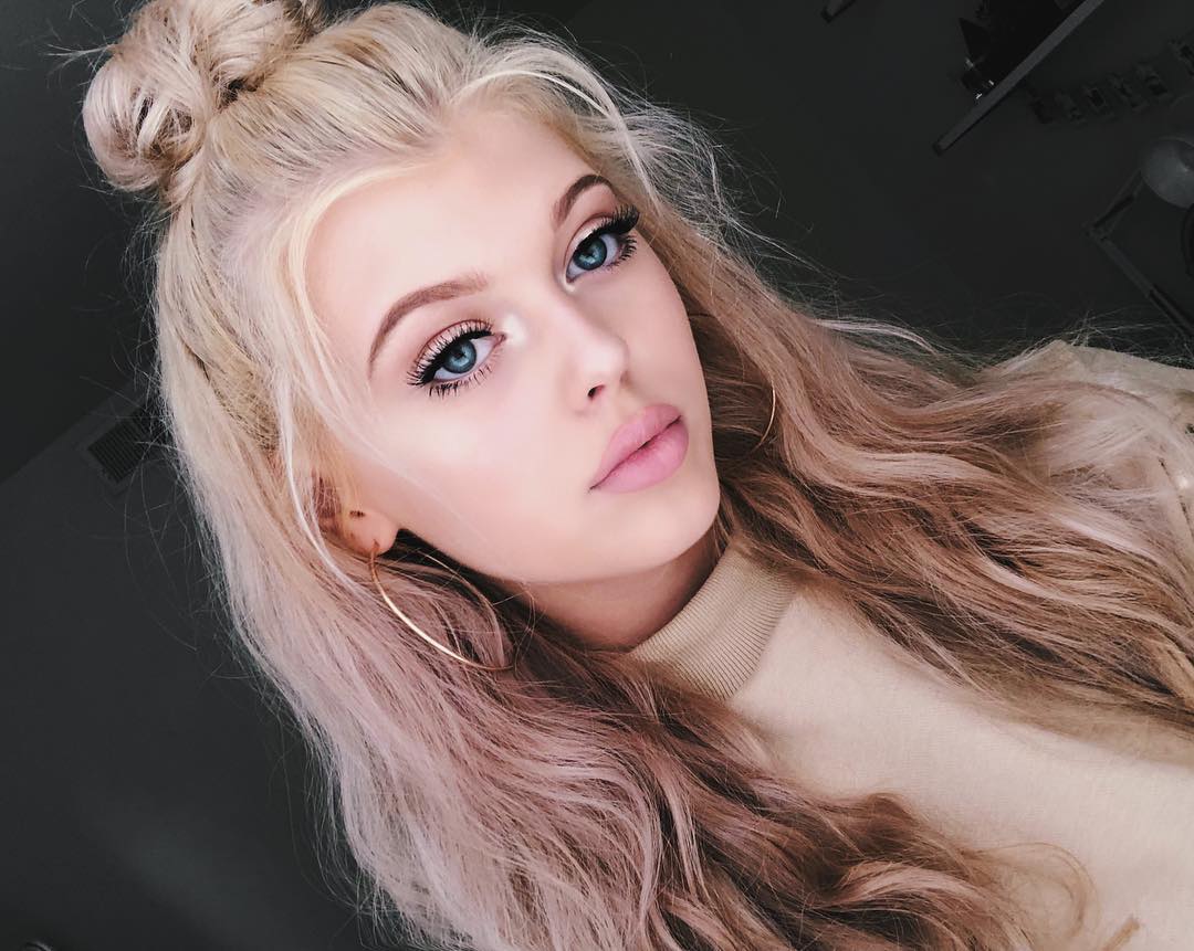a closeup portrait of a young woman with blonde hair styled in a bun and loose waves. she has bright blue eyes and is wearing makeup, including eyeliner, mascara, and light pink lipstick. she is wearing gold hoop earrings and a light beige turtleneck sweater. the background is a dark gray color. she is looking directly at the camera with a subtle expression.