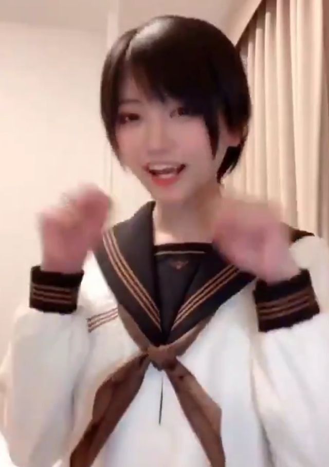 a young woman with short black hair is wearing a white sailor uniform with a black collar and a brown ribbon. she is smiling and looking at the camera. she is standing in a room with a white wall and a brown curtain in the background. she has a youthful appearance and her expression suggests she is happy and energetic.