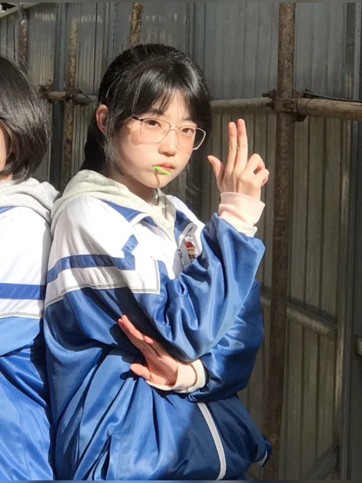 a young asian girl in a blue and white school uniform and eyeglasses is posing outdoors. she has a lollipop in her mouth and is making a hand gesture with her index and middle fingers pointed up, like a gun. she has bangs and her hair is pulled back. she is standing next to another person in a similar uniform, with a metal fence behind them. sunlight creates a contrast of light and shadow.