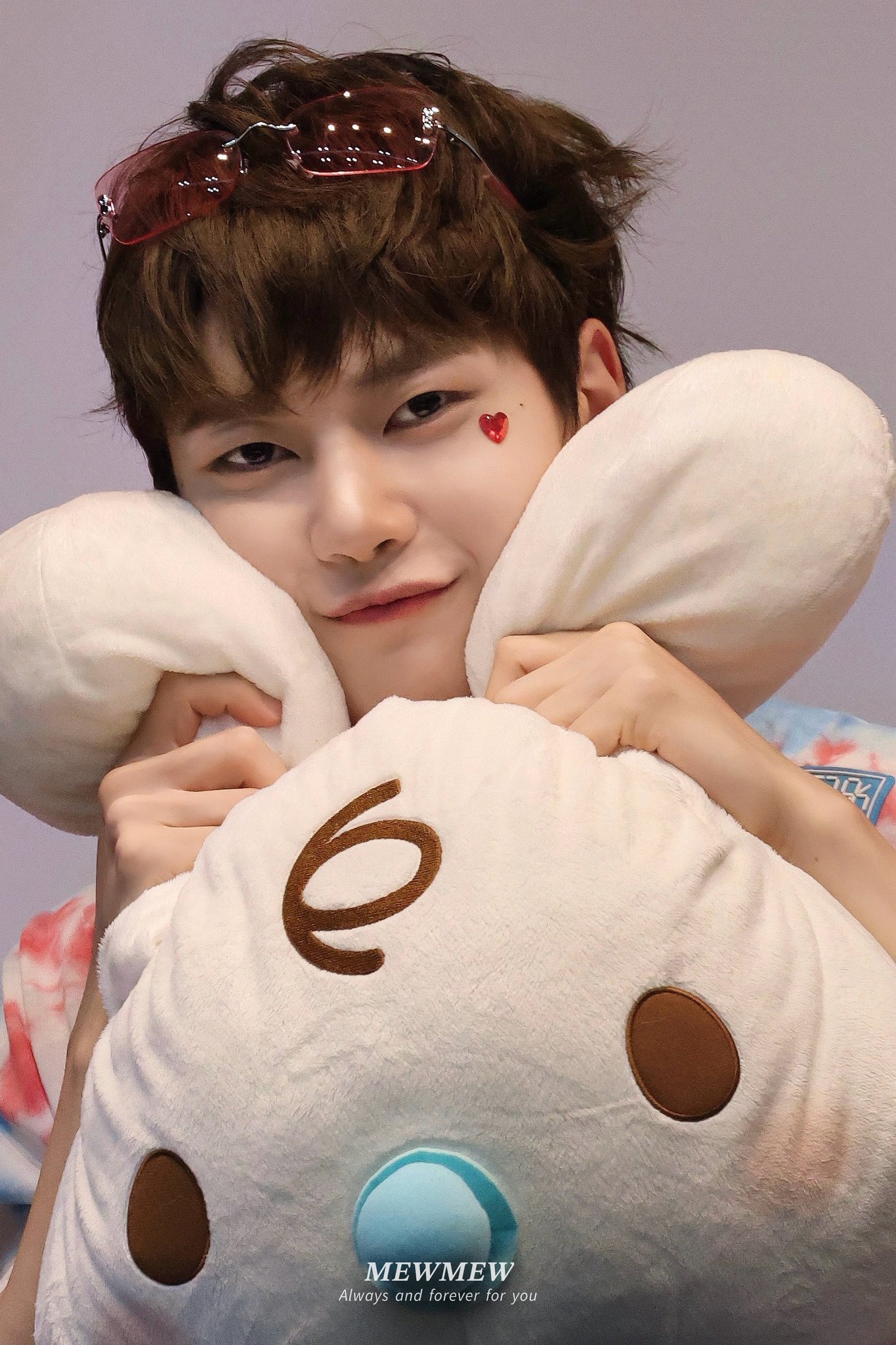 a young asian man smiles softly at the camera while hugging a large white plush toy with brown accents. he wears a pink heart sticker below his eye and a tiedye shirt in shades of pink and blue. the photo appears to be taken indoors with soft, artificial lighting.