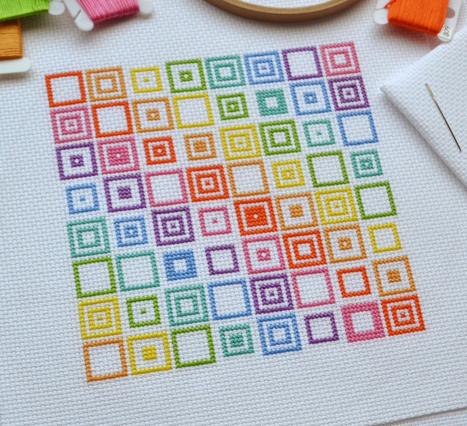a closeup shot of a crossstitch project. the project is a rainbow of squares, each progressively getting larger as they move from one side to the other. the background is a white fabric.  the stitching is neat and even, and the colors are vibrant. the project appears to be nearly finished.