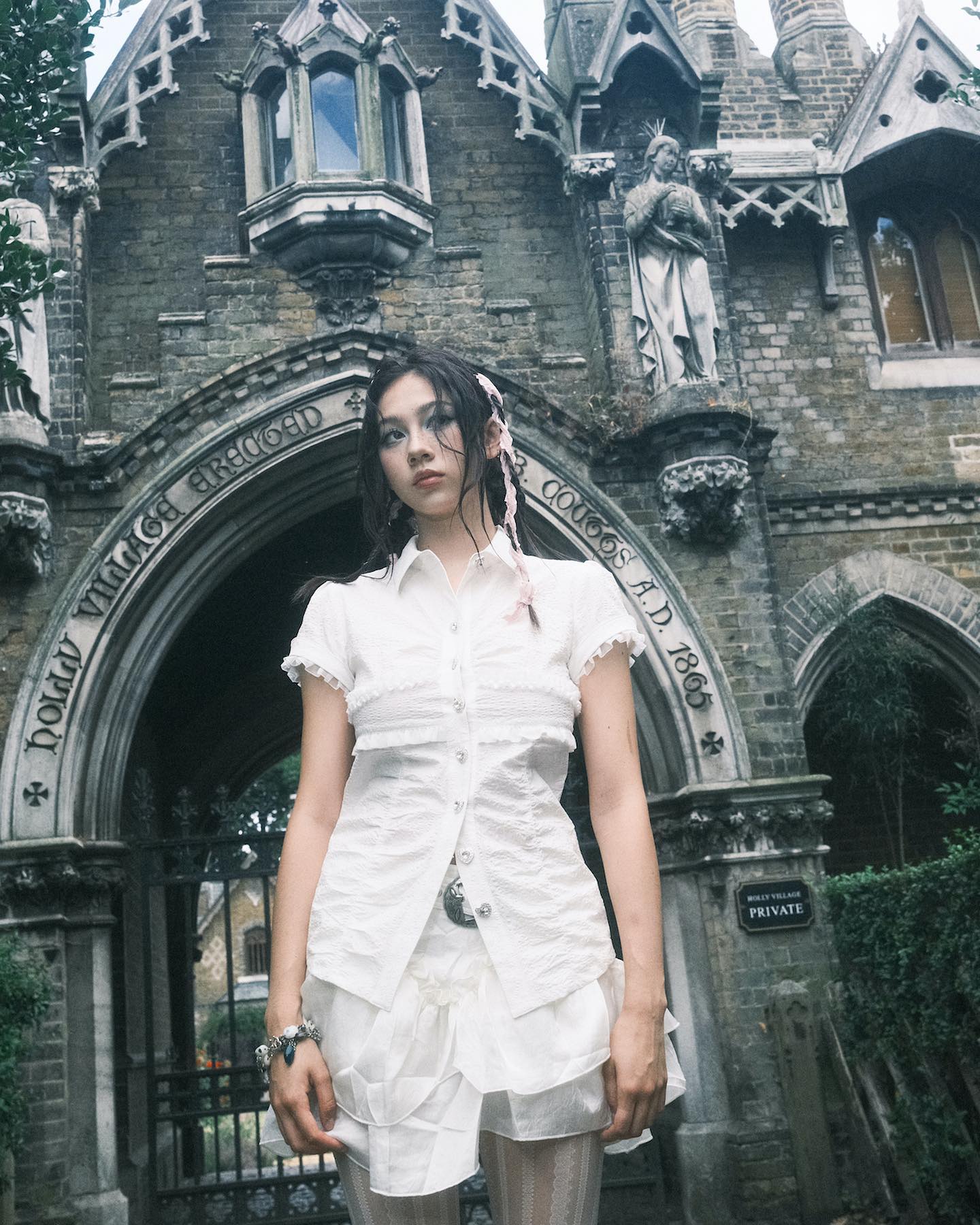 young woman in a white ruffled shirt and pleated skirt stands in an archway of an old gothic style building. the building is made of grey stone and has intricate carvings and statues. the woman's dress is reminiscent of vintage or gothic fashion. the overall aesthetic is dark, moody, and slightly vintage, possibly due to the film photography style.