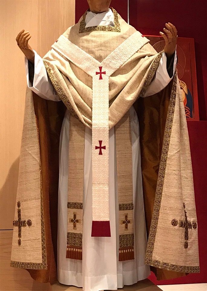 a priest in a white robe with a gold cape. he is wearing a white stole with red crosses. the priest is standing in front of a red wall. the priest's face is not visible. the photo is taken from the front.
